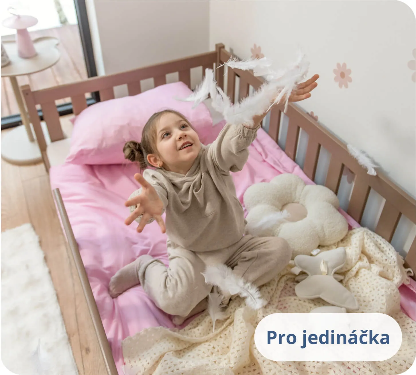 Holčička hrající si s bílými pírky na růžovém povlečení v posteli čokoládové barvy.